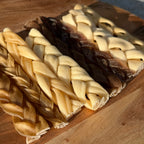 Two braided dog chew treats, one pale cream and one dark brown, arranged on a wooden cutting board.