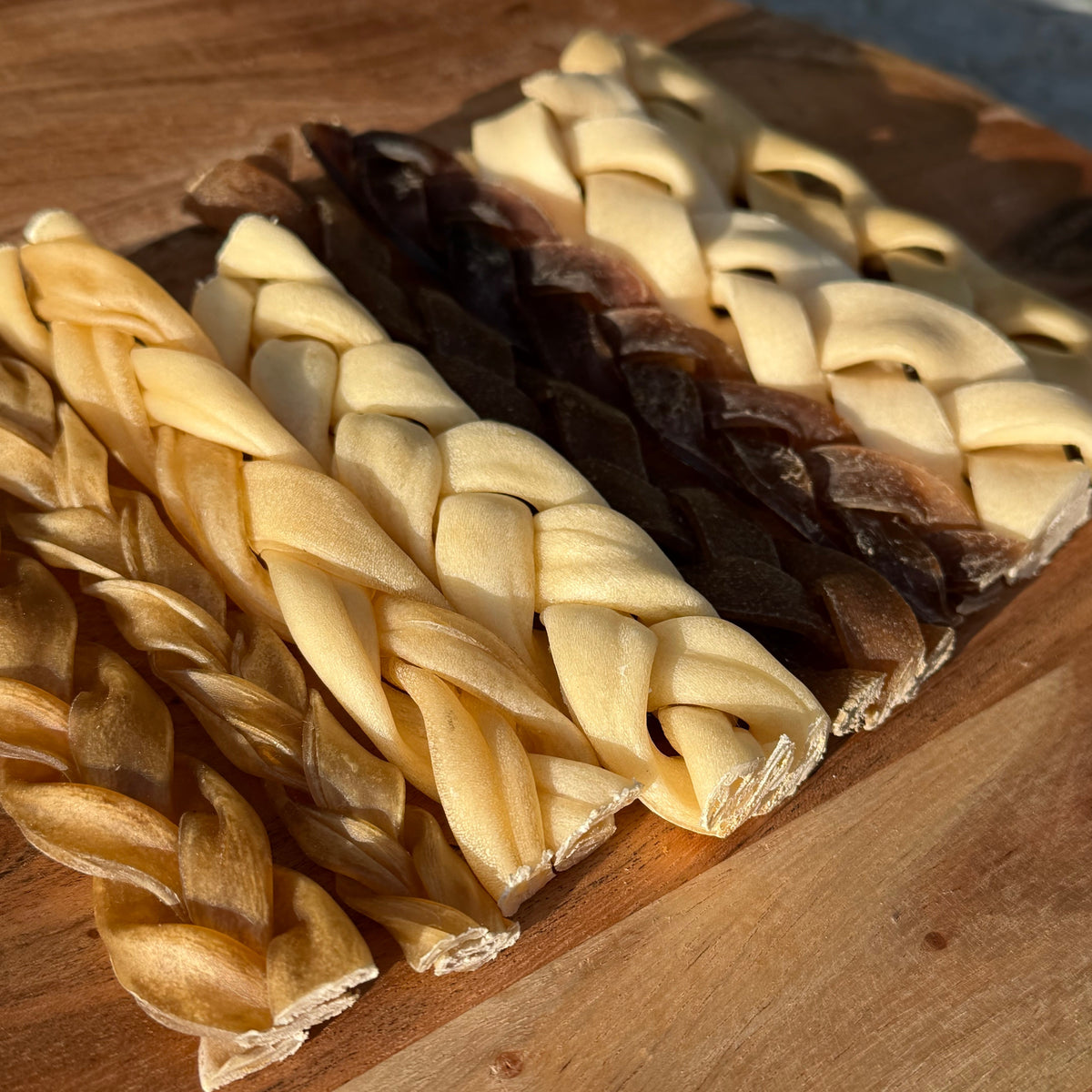 Two braided dog chew treats, one pale cream and one dark brown, arranged on a wooden cutting board.